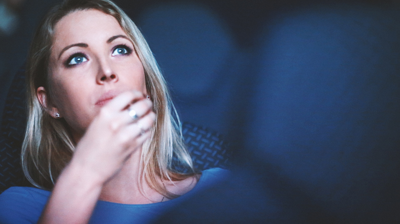 Closeup of mid 20's blond woman watching a movie at cinema place and eating popcorn. Low angle view, shot between seats in front.